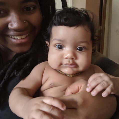 African American Mom Holding A Teething Baby Wearing Amber Guru's Raw Unpolished Baltic Amber Teething Necklace For Babies In Green Color