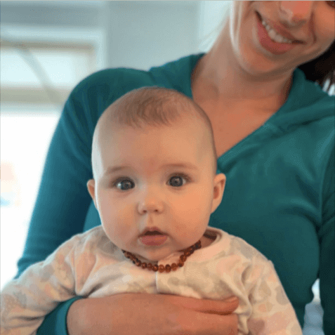 Mom Holding Little Teething Baby Girl Wearing Amber Guru's Polished Baltic Amber Teething Necklace For Children In Cognac Color