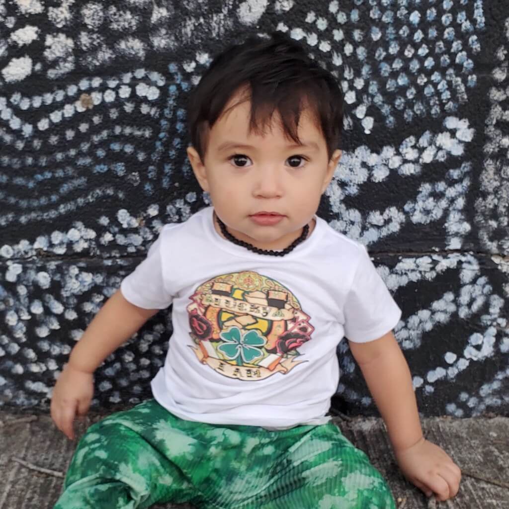 Baby Boy with Dark Hair and Eyes Wearing Raw Black Baltic Amber Teething Necklace, White T-shirt and Green Pants, Sitting on the Floor