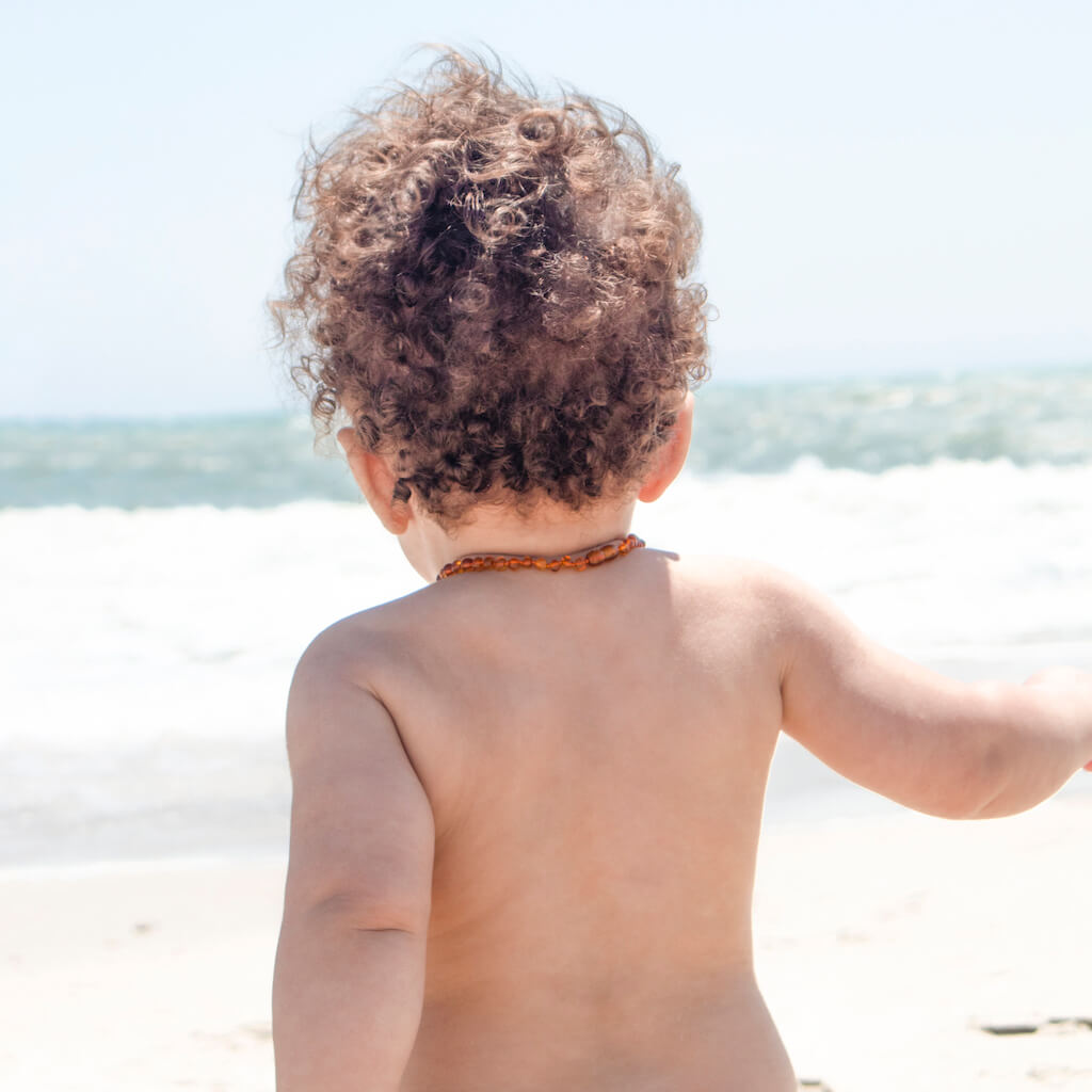 Curly-haired Teething Baby Boy with Baltic Amber Necklace Walking on the Beach Towards the Baltic Sea