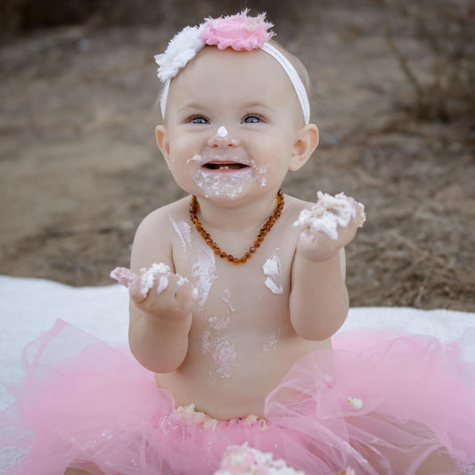 Happy Teething Baby Girl with Birthday Cake Frosting, Wearing Amber Guru Cognac Color Baltic Amber Teething Necklace with Polished Beads