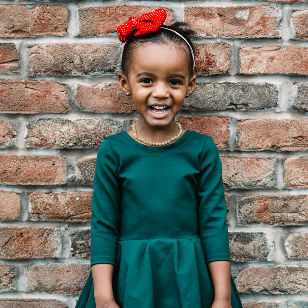 Smiling Girl with Amber Guru Yellow Baltic Amber Necklace, Red Bow and Green Dress, Standing by the Wall