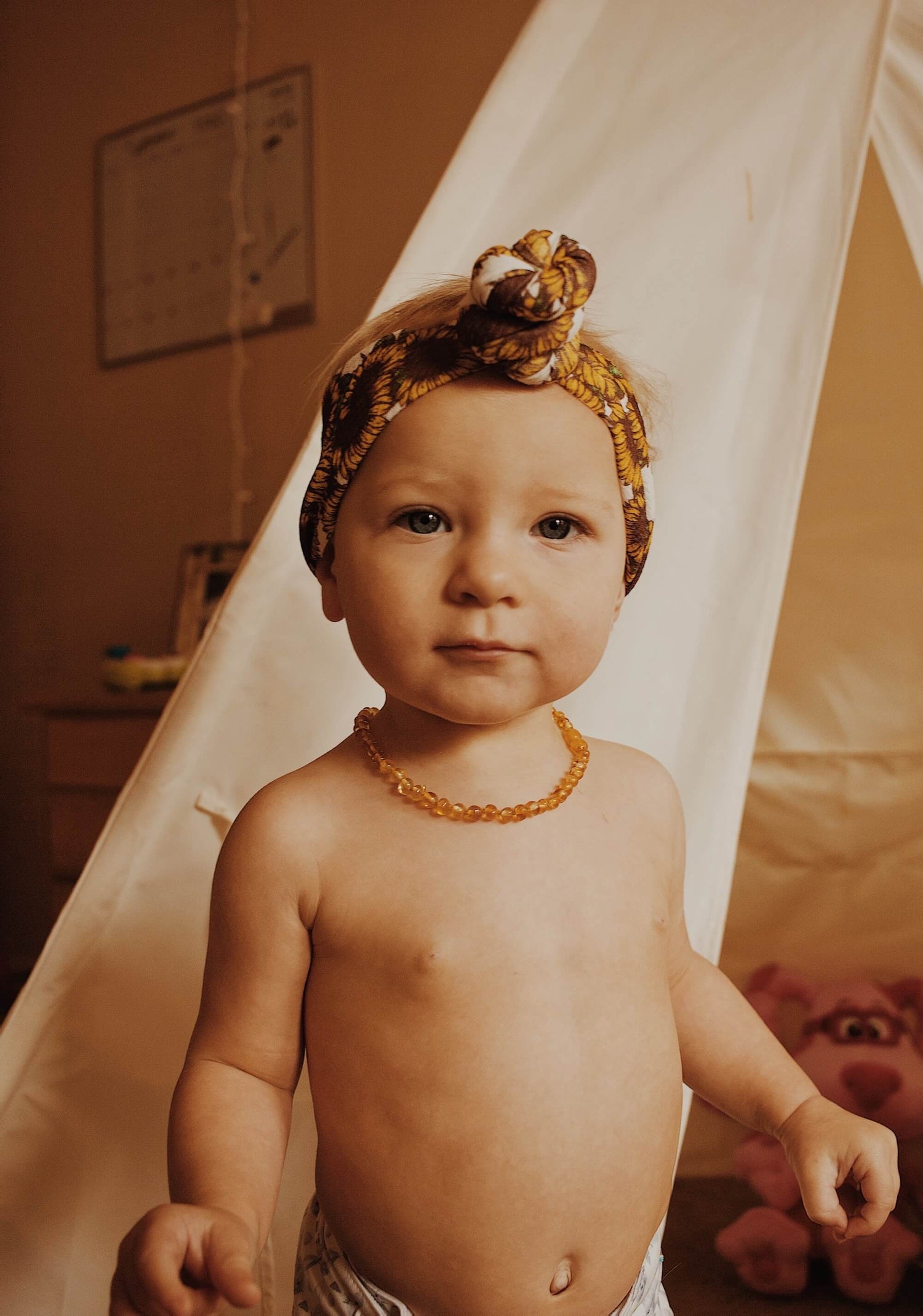 Baby Girl Wearing Amber Guru's Baltic Amber Teething Necklace In Honey Color, Posing In A Room