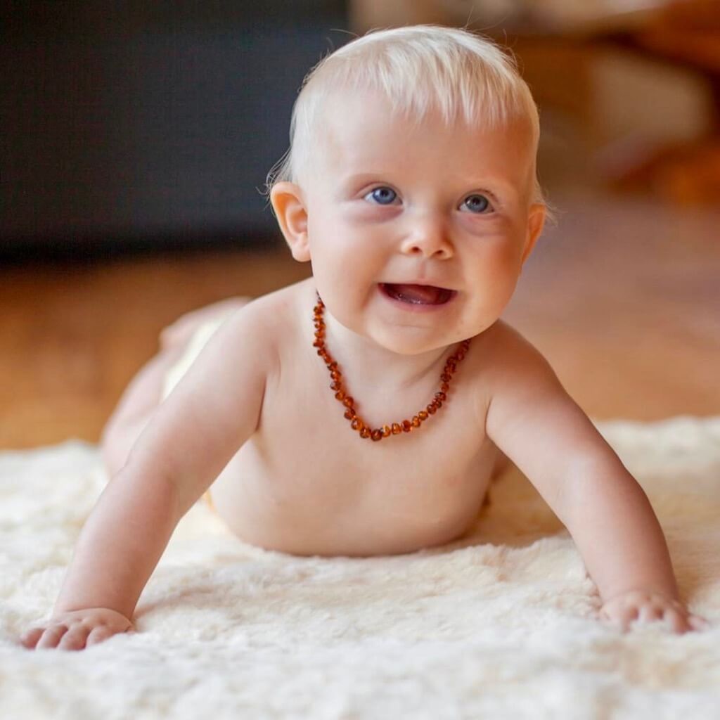 Blond-haired and Blue-eyed Baby Boy, Crawling on a White Carpet, Wearing Amber Teething Necklace Made from 100% Baltic Amber, in Cognac Color, with Polished Beads