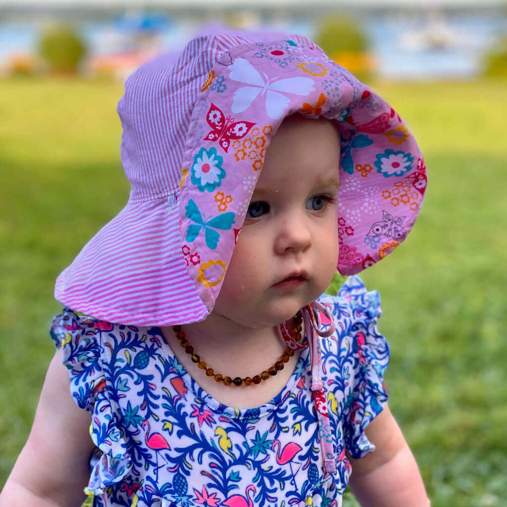 Cute Baby Girl, Wearing Pink Hat and Baltic Amber Teething Necklace in Cognac Color, Walking on Green Grass