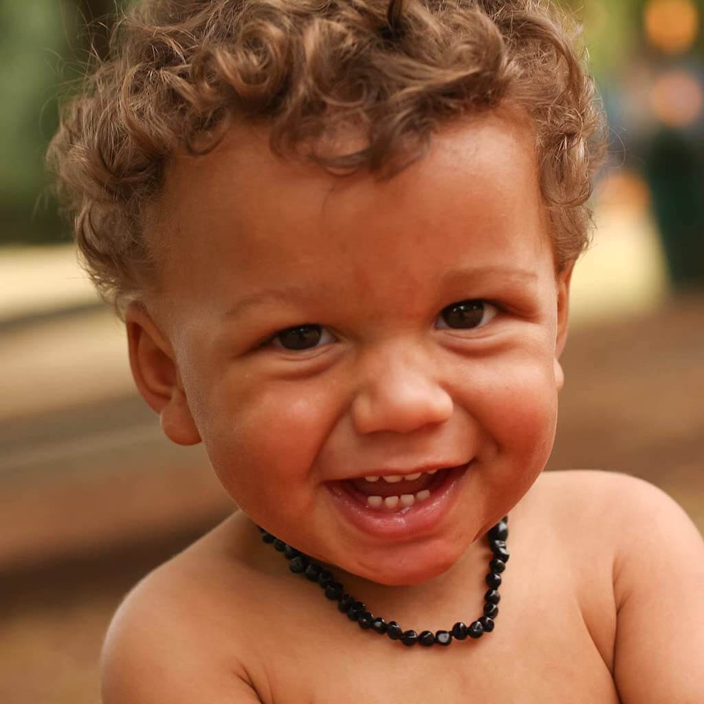 Smiling Baby Boy with Brown Curly Hair and Dark Eyes, Wearing Amber Guru Baltic Amber Teething Necklace with Polished Beads in Cherry Color