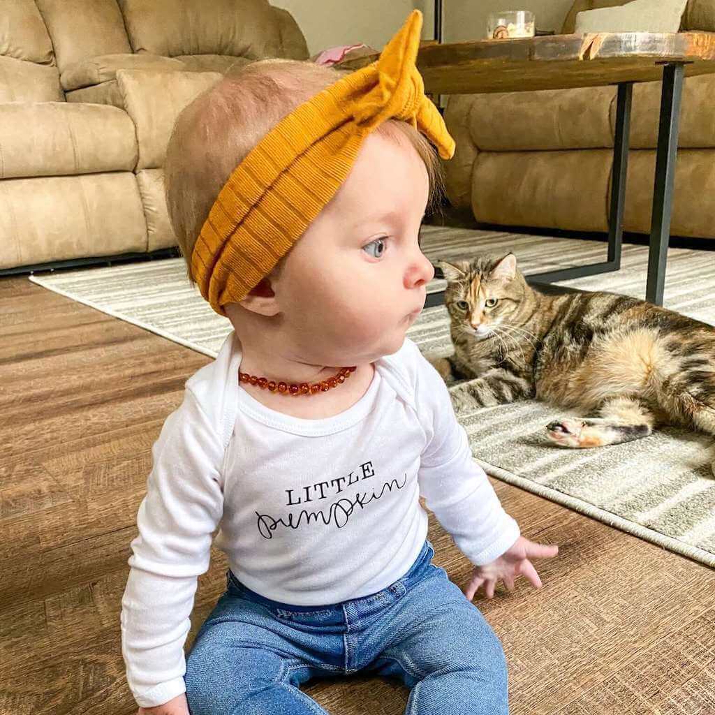 Surprised Baby Girl Wearing Premium Cognac Amber Teething Necklace from Amber Guru, and Headband, with Cat in the Room at Home