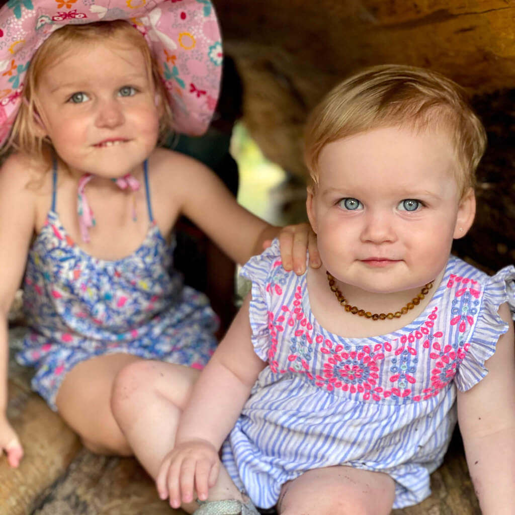 Two Blue-eyed Baby Girls Sitting Inside the Tree and One of Them is Wearing Premium Amber Teething Necklace with Raw Green Beads from Amber Guru